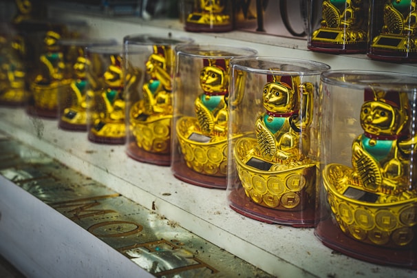 A row of gold-colored waving cat figurines enclosed in transparent cases is displayed on a shelf. Each cat is adorned with a red collar and stands on a base resembling a golden boat filled with coins. The background includes reflections and a dimly lit store ambiance.