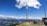 A person is swinging high in the sky on a large swing suspended between two tall poles. Below, a group of people are gathered, some sitting at a picnic table and others standing, enjoying the panoramic view. The background features a stunning landscape of mountains and valleys under a clear blue sky with scattered clouds.