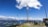 A person is swinging high in the sky on a large swing suspended between two tall poles. Below, a group of people are gathered, some sitting at a picnic table and others standing, enjoying the panoramic view. The background features a stunning landscape of mountains and valleys under a clear blue sky with scattered clouds.