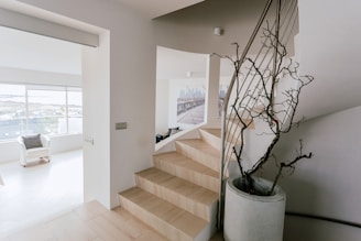 An interior space featuring a modern staircase, a large ceramic pot with a decorative, barren branch, and an open, bright living area with a white armchair near a window. The room has light-colored floors and walls, creating a minimalist and airy atmosphere.