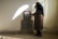 A person wearing a dark headscarf stands in a somber pose, placing a hand on a framed photograph set atop a stone marker. The room is dimly lit with sunlight filtering through blinds, casting shadows across the walls and floor.