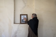 An elderly person with gray hair wearing a dark shawl stands in front of a plain, textured wall. They are touching a framed photograph of a man in a suit that is placed on a small ledge, creating a sense of nostalgia or remembrance.