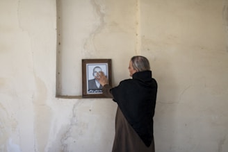 An elderly person with gray hair wearing a dark shawl stands in front of a plain, textured wall. They are touching a framed photograph of a man in a suit that is placed on a small ledge, creating a sense of nostalgia or remembrance.
