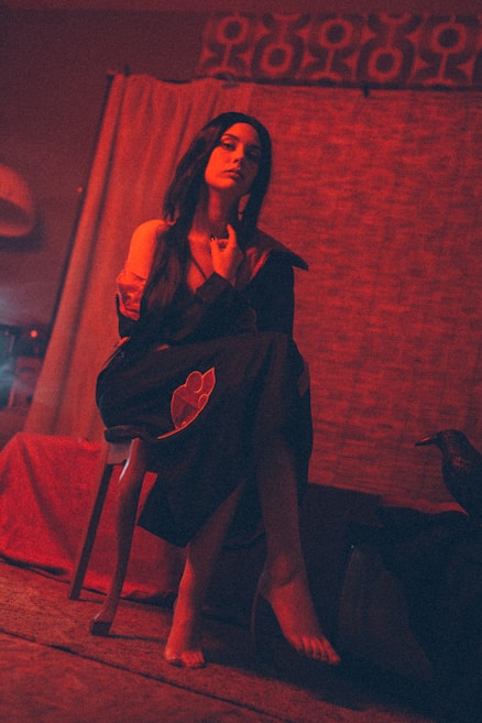 A moody, dimly lit setting with a person sitting on a chair, draped in a dark robe with red detailing. The ambiance appears intimate and slightly mysterious, with warm, reddish lighting casting shadows. A decorative curtain and patterned wall textile add depth to the background, and a raven-like sculpture is visible to the side.
