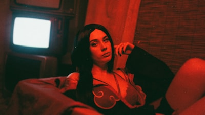 A woman with dark hair is lounging on a couch, illuminated by a red light, creating a somber yet intriguing atmosphere. A vintage television is visible in the background, casting a bright glow.