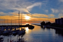 A picturesque sunset over a marina, with boats docked along the calm water reflecting the vibrant colors of the sky. The silhouette of buildings and trees outline the horizon, while streaks of orange, yellow, and blue paint the sky.