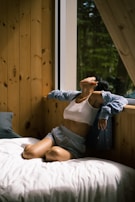 A person in casual clothing, including a white tank top, shorts, and an open cardigan, is sitting on a bed next to a large window. The person appears to be in a relaxed pose with their head tilted back and eyes closed. The room has wooden walls and there is a forest view outside the window.