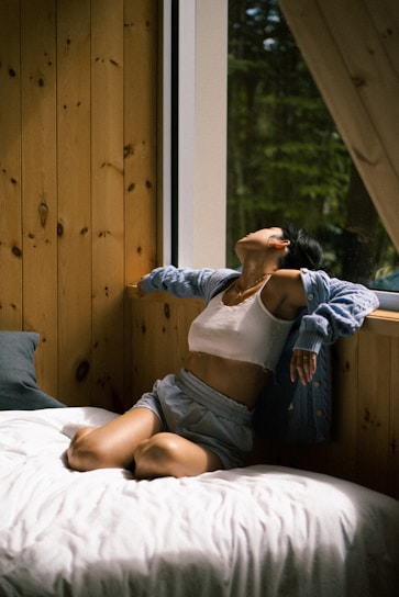 A person in casual clothing, including a white tank top, shorts, and an open cardigan, is sitting on a bed next to a large window. The person appears to be in a relaxed pose with their head tilted back and eyes closed. The room has wooden walls and there is a forest view outside the window.