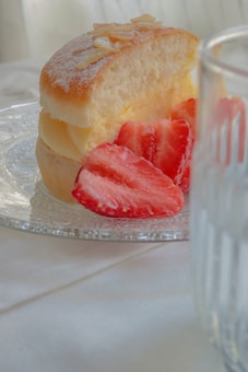 A soft, fluffy bun dusted with powdered sugar is filled with cream and topped with thin almond slices. Freshly cut strawberries are placed beside the bun, resting on a clear, textured glass plate. A glass with a linear pattern is partially visible in the foreground, creating a gentle, blurred effect.