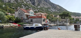 A tranquil coastal scene with a stone house featuring red-tiled roofs, set against a backdrop of lush green hills. A small boat is moored by the water next to a dock, with calm waters reflecting the clear sky.