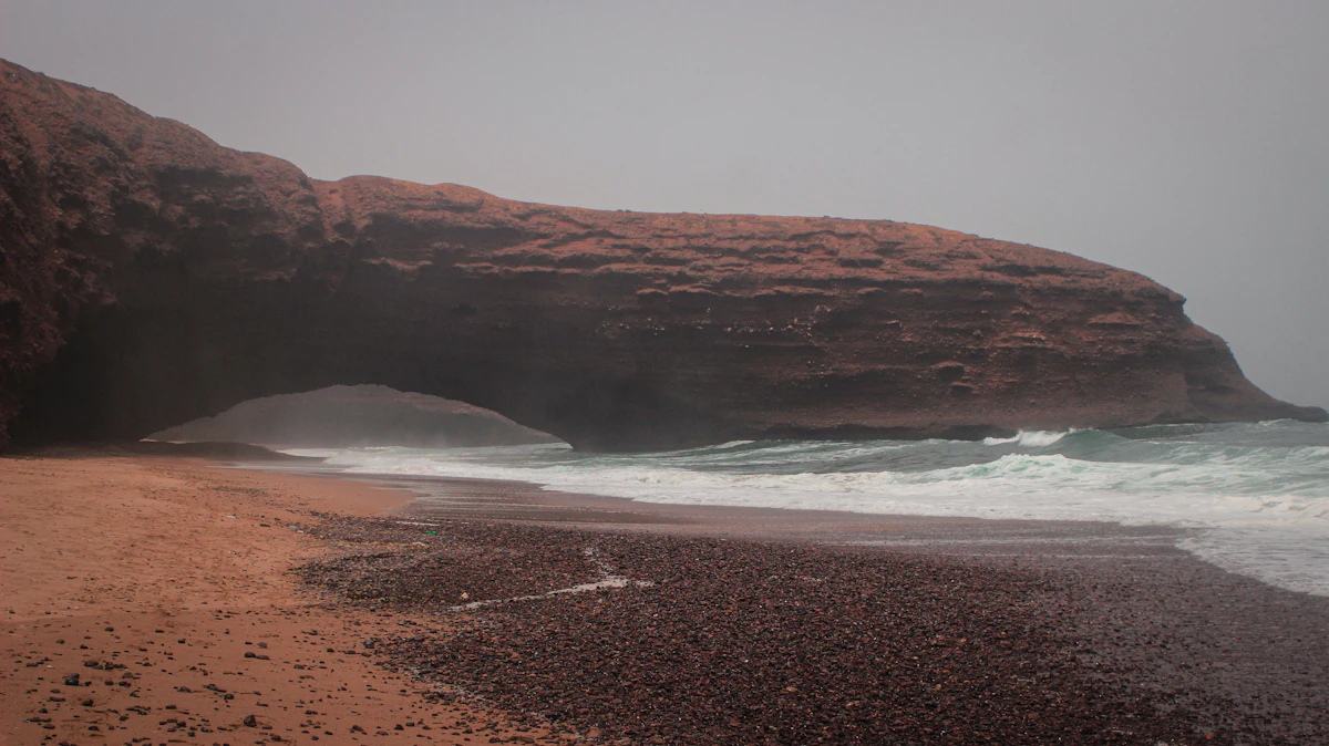 Grey surf below a long red cliff and arch on a misty day at Legzira