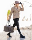 A person walks confidently, dressed in a beige leather jacket, black ripped jeans, and chunky black boots. They wear a leopard print hat and black gloves, accessorized with sunglasses and a gold chain necklace. The person carries a designer travel bag in one hand.