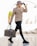 A person walks confidently, dressed in a beige leather jacket, black ripped jeans, and chunky black boots. They wear a leopard print hat and black gloves, accessorized with sunglasses and a gold chain necklace. The person carries a designer travel bag in one hand.