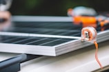 A close-up view of a measuring tape placed on the edge of a solar panel. The focus is on the transparent measuring tape with an orange strap, while the solar panel beneath has a metallic frame. The background is slightly out of focus, adding depth to the composition.