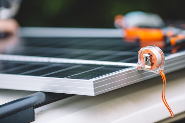A close-up view of a measuring tape placed on the edge of a solar panel. The focus is on the transparent measuring tape with an orange strap, while the solar panel beneath has a metallic frame. The background is slightly out of focus, adding depth to the composition.