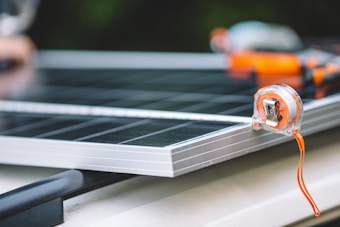 A close-up view of a measuring tape placed on the edge of a solar panel. The focus is on the transparent measuring tape with an orange strap, while the solar panel beneath has a metallic frame. The background is slightly out of focus, adding depth to the composition.