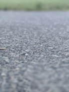 A close-up view of an asphalt surface, with small rocks and pebbles visible. The background is blurred, suggesting an outdoor setting possibly near a field.