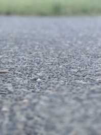A close-up view of an asphalt surface, with small rocks and pebbles visible. The background is blurred, suggesting an outdoor setting possibly near a field.