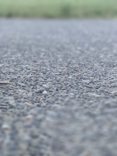 A close-up view of an asphalt surface, with small rocks and pebbles visible. The background is blurred, suggesting an outdoor setting possibly near a field.