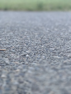 A close-up view of an asphalt surface, with small rocks and pebbles visible. The background is blurred, suggesting an outdoor setting possibly near a field.