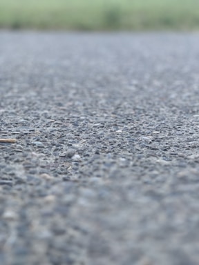 A close-up view of an asphalt surface, with small rocks and pebbles visible. The background is blurred, suggesting an outdoor setting possibly near a field.