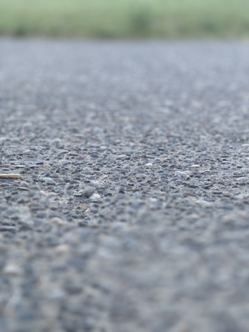 A close-up view of an asphalt surface, with small rocks and pebbles visible. The background is blurred, suggesting an outdoor setting possibly near a field.