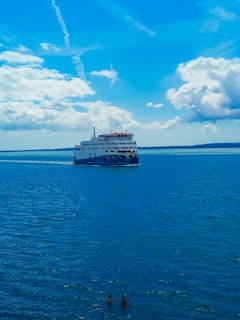 A large ferry is traveling across a vast expanse of blue water under a sky filled with fluffy, scattered clouds. Two people are swimming in the foreground. The scene captures a sense of travel and leisure on a bright, sunny day.