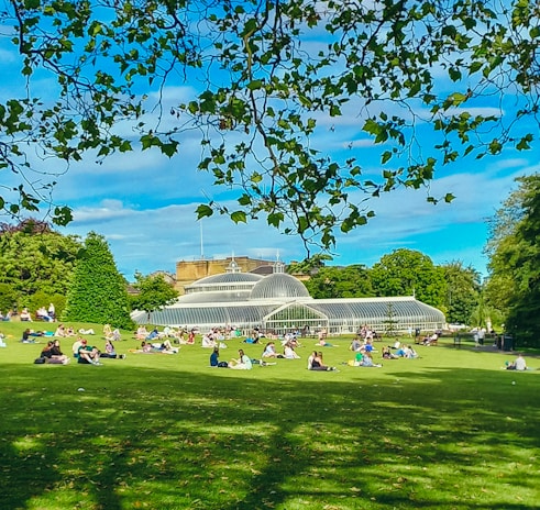 A large green lawn filled with people relaxing and enjoying leisure time sits in front of a grand glass greenhouse structure. The sky above is a vibrant blue, and leafy branches frame the upper part of the scene. The area is surrounded by lush trees and greenery.