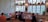 A cozy café interior with wooden furniture, colorful cushions, and vintage decor on the walls. People are seated at tables, enjoying their meals and drinks. The large windows allow natural light to brighten the room, with potted plants adorning the ledges.