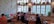 A cozy café interior with wooden furniture, colorful cushions, and vintage decor on the walls. People are seated at tables, enjoying their meals and drinks. The large windows allow natural light to brighten the room, with potted plants adorning the ledges.