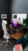 A modern and organized home office setup with a minimalist design. A wooden desk holds a computer monitor displaying a gradient screen, a keyboard, and a mouse. The desk is accessorized with small plants, a speaker, and decorative items. A white ergonomic office chair stands in front of the desk. The wall behind the desk features a pegboard with hanging items and more plants, and another wall displays a framed photo or artwork. The area also includes a basket with a book titled 'MOC' inside.
