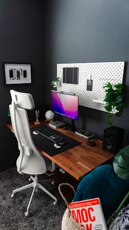 A modern and organized home office setup with a minimalist design. A wooden desk holds a computer monitor displaying a gradient screen, a keyboard, and a mouse. The desk is accessorized with small plants, a speaker, and decorative items. A white ergonomic office chair stands in front of the desk. The wall behind the desk features a pegboard with hanging items and more plants, and another wall displays a framed photo or artwork. The area also includes a basket with a book titled 'MOC' inside.