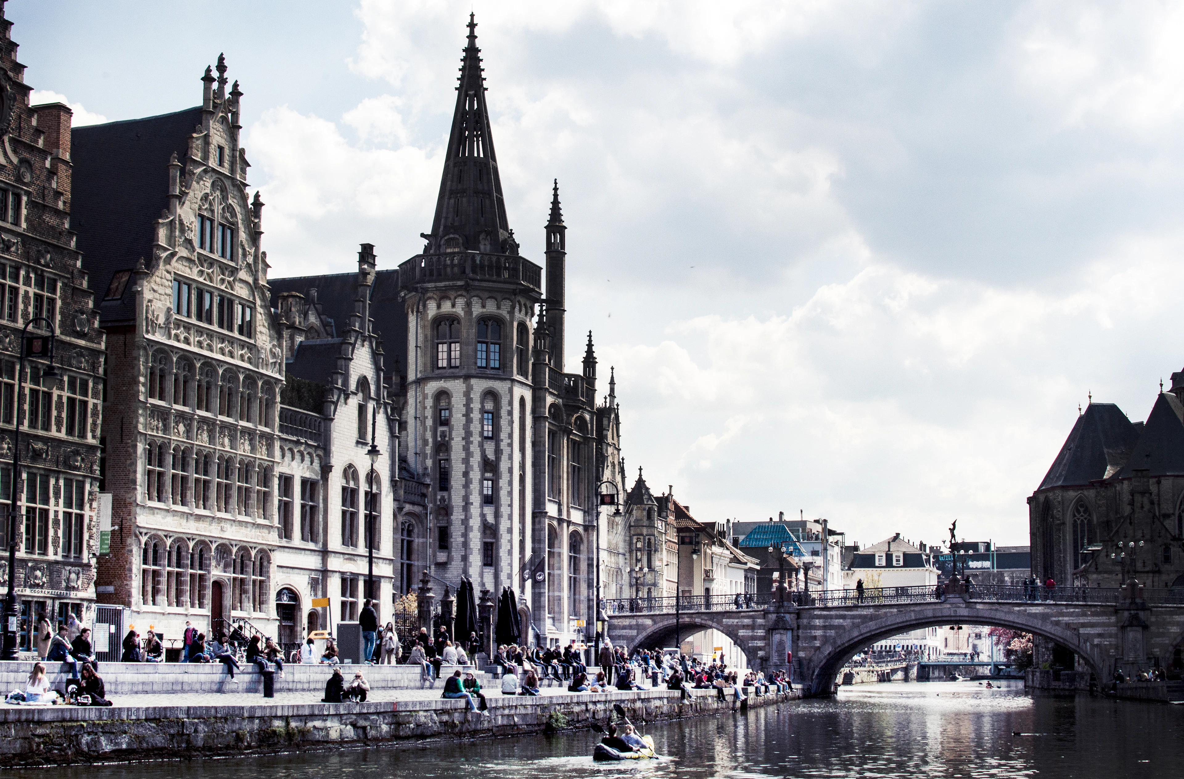 Gothic guild houses along Ghent's Graslei quay in warm afternoon light, with people relaxing by the water and a bridge beyond.