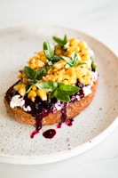 A slice of toasted bread topped with creamy cheese, sweet corn kernels, fresh basil leaves, and a drizzle of dark berry sauce on a speckled ceramic plate.