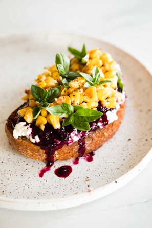 A slice of toasted bread topped with creamy cheese, sweet corn kernels, fresh basil leaves, and a drizzle of dark berry sauce on a speckled ceramic plate.