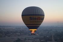 A hot air balloon floats serenely over a cityscape during the early morning hours. The sky is a soft gradient of light colors as the sun begins to rise, providing a tranquil backdrop. The balloon, featuring the logo 'Sindbad', is gently illuminated from below by the burner flame, and the landscape below is shrouded in a light mist.