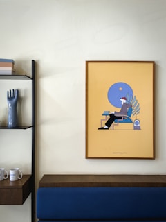 A minimalist interior featuring a framed piece of art with abstract design hanging on a light-colored wall. The art has a yellow background with blue and dark tones. Next to it is a modern shelf with a glossy, dark vase shaped like a hand and several stacked books. Below the art and shelf, a wooden ledge and a blue upholstered bench are visible.