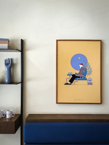 A minimalist interior featuring a framed piece of art with abstract design hanging on a light-colored wall. The art has a yellow background with blue and dark tones. Next to it is a modern shelf with a glossy, dark vase shaped like a hand and several stacked books. Below the art and shelf, a wooden ledge and a blue upholstered bench are visible.