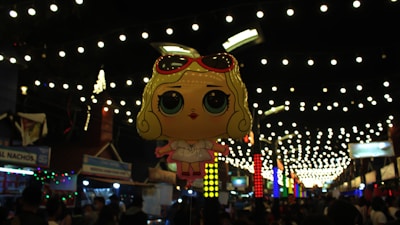 A cartoonish balloon figure depicting a character with large eyes and blonde hair under a canopy of festive string lights. The balloon hovers above a busy street market with colorful stalls and signs, creating an energetic and vibrant atmosphere.
