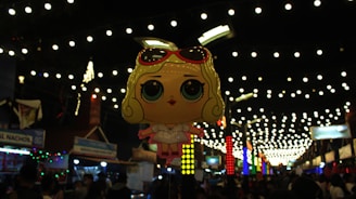 A cartoonish balloon figure depicting a character with large eyes and blonde hair under a canopy of festive string lights. The balloon hovers above a busy street market with colorful stalls and signs, creating an energetic and vibrant atmosphere.