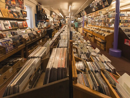 A record store with rows of vinyl records and CDs neatly arranged. The store is filled with shelves displaying album covers, and there are a few people browsing through the collection.