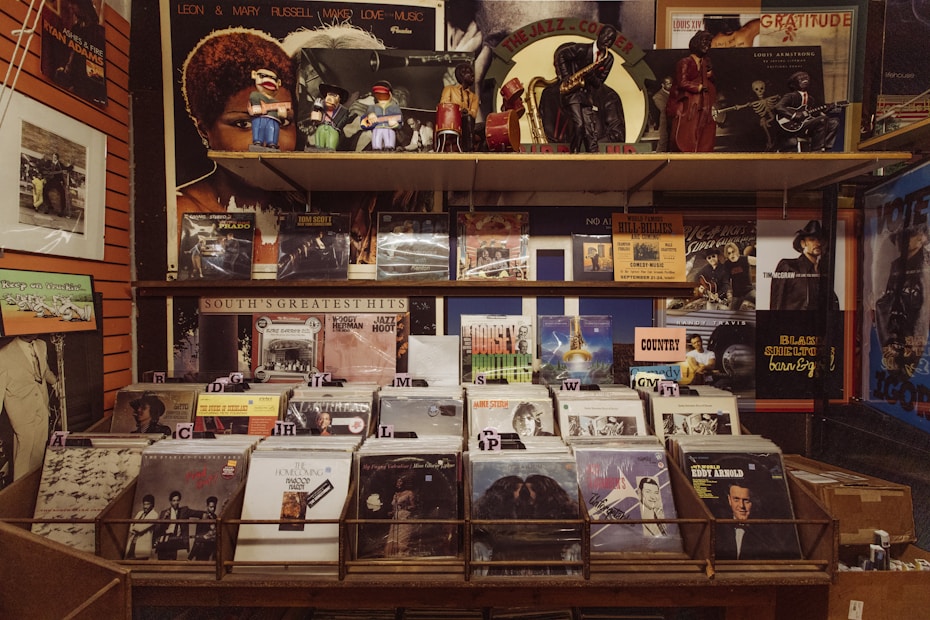 A vintage record store display features a wide array of vinyl records sorted into sections, with prominent labels. Above the records, retro figurines and posters of jazz musicians and album covers are displayed, creating a nostalgic atmosphere.