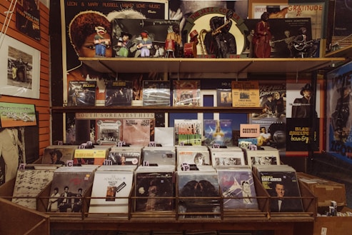 A vintage record store display features a wide array of vinyl records sorted into sections, with prominent labels. Above the records, retro figurines and posters of jazz musicians and album covers are displayed, creating a nostalgic atmosphere.