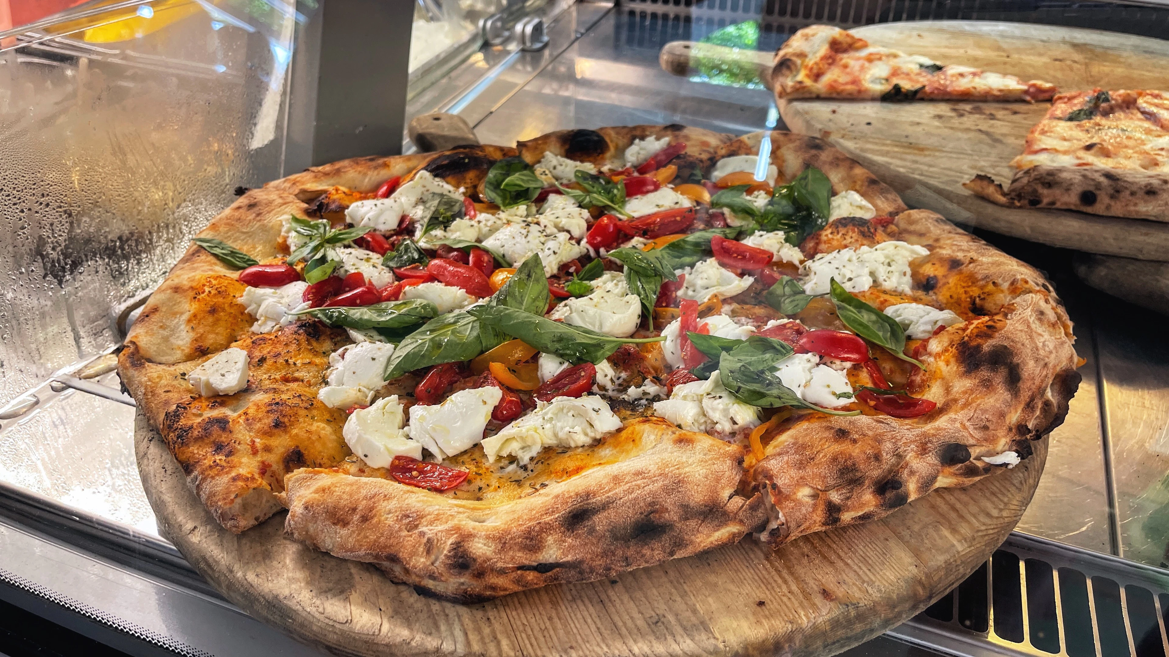 A fresh Margherita pizza with mozzarella and basil sits on a wooden board in a pizzeria display.
