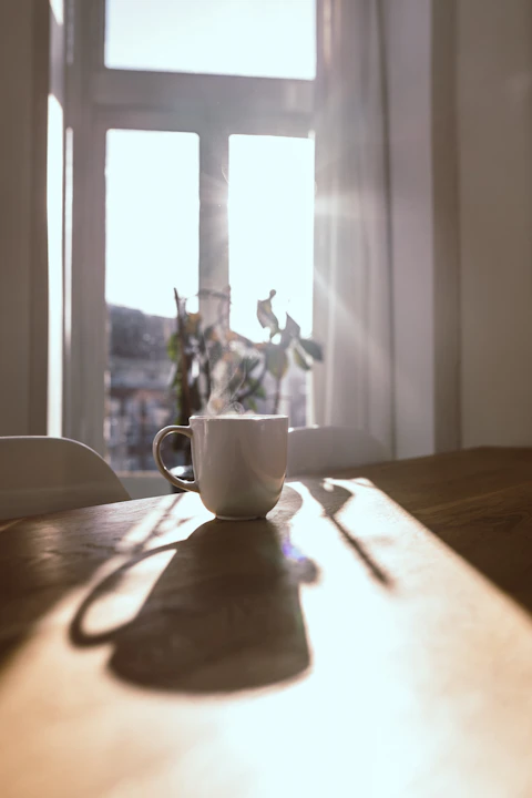 A steaming white mug on a wooden table in front of a sunlit window