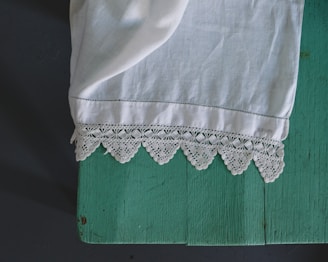 A piece of white fabric with intricate lace detailing lies on a teal wooden surface. The fabric appears to be elegantly draped, with the lace edges prominently displayed.