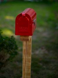 A red mailbox with 'U.S. Mail' written on it is mounted on a wooden post. It is situated in a grassy area with some green foliage nearby.
