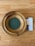A wooden offering plate with a green felt base is placed on a light wooden floor. Next to the plate, there is a pamphlet partially visible, featuring text and images related to church offerings.