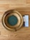 A wooden offering plate with a green felt base is placed on a light wooden floor. Next to the plate, there is a pamphlet partially visible, featuring text and images related to church offerings.