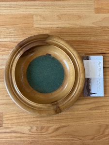 A wooden offering plate with a green felt base is placed on a light wooden floor. Next to the plate, there is a pamphlet partially visible, featuring text and images related to church offerings.